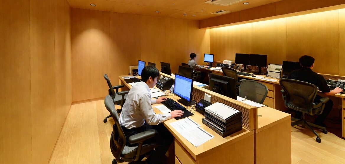 木製の壁と天井のモダンなオフィスで、パソコンが置かれた机に向かって仕事をする3人。机の上には書類や事務用品が置かれ、頭上と壁面の照明で部屋は明るい。.